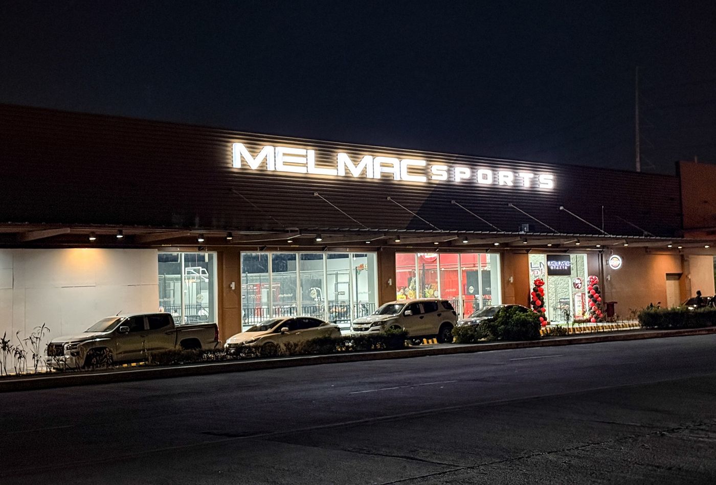 The lit Melmac Sports building at Vista Mall Imus at night, seen from the car park
