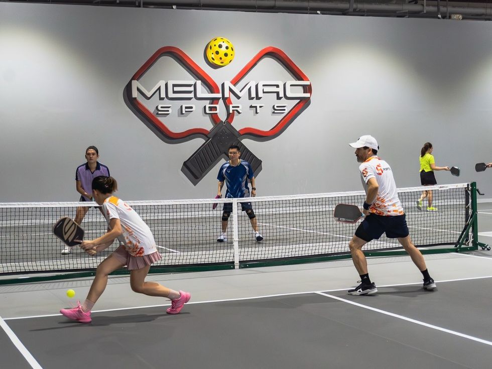 A doubles pickleball rally under the Melmac paddle wall mark at Imus