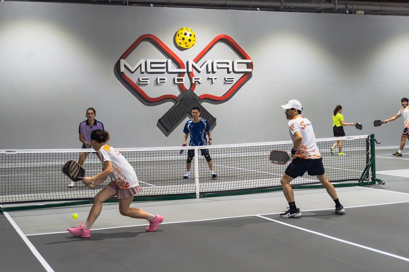A doubles pickleball rally under the Melmac paddle wall mark at Imus