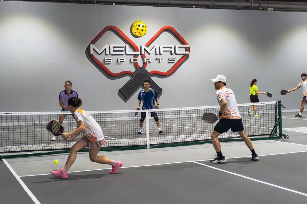 A doubles pickleball rally under the Melmac paddle wall mark at Imus