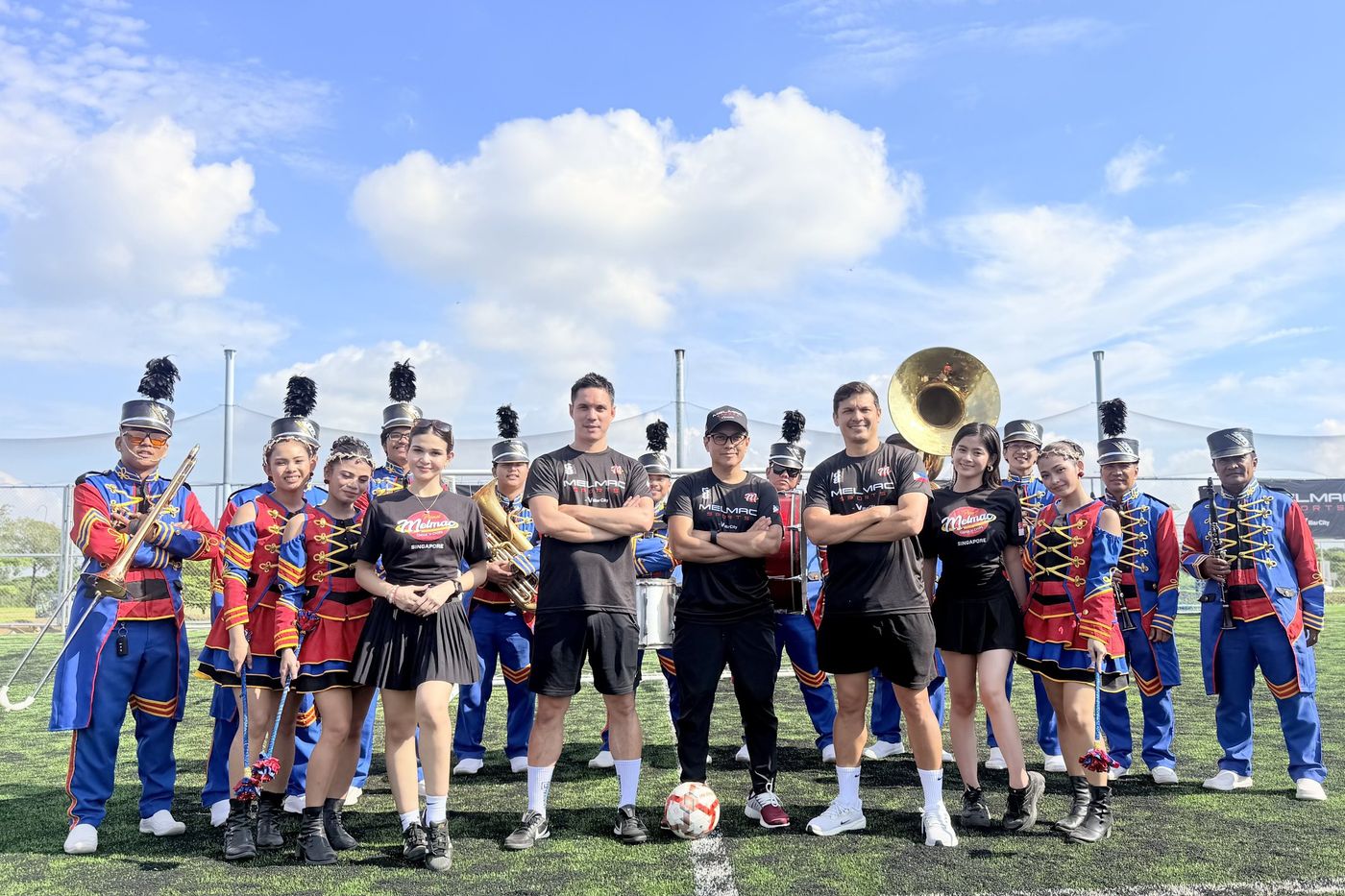 The marching band and players in black Melmac shirts posing together on the pitch at Villar City