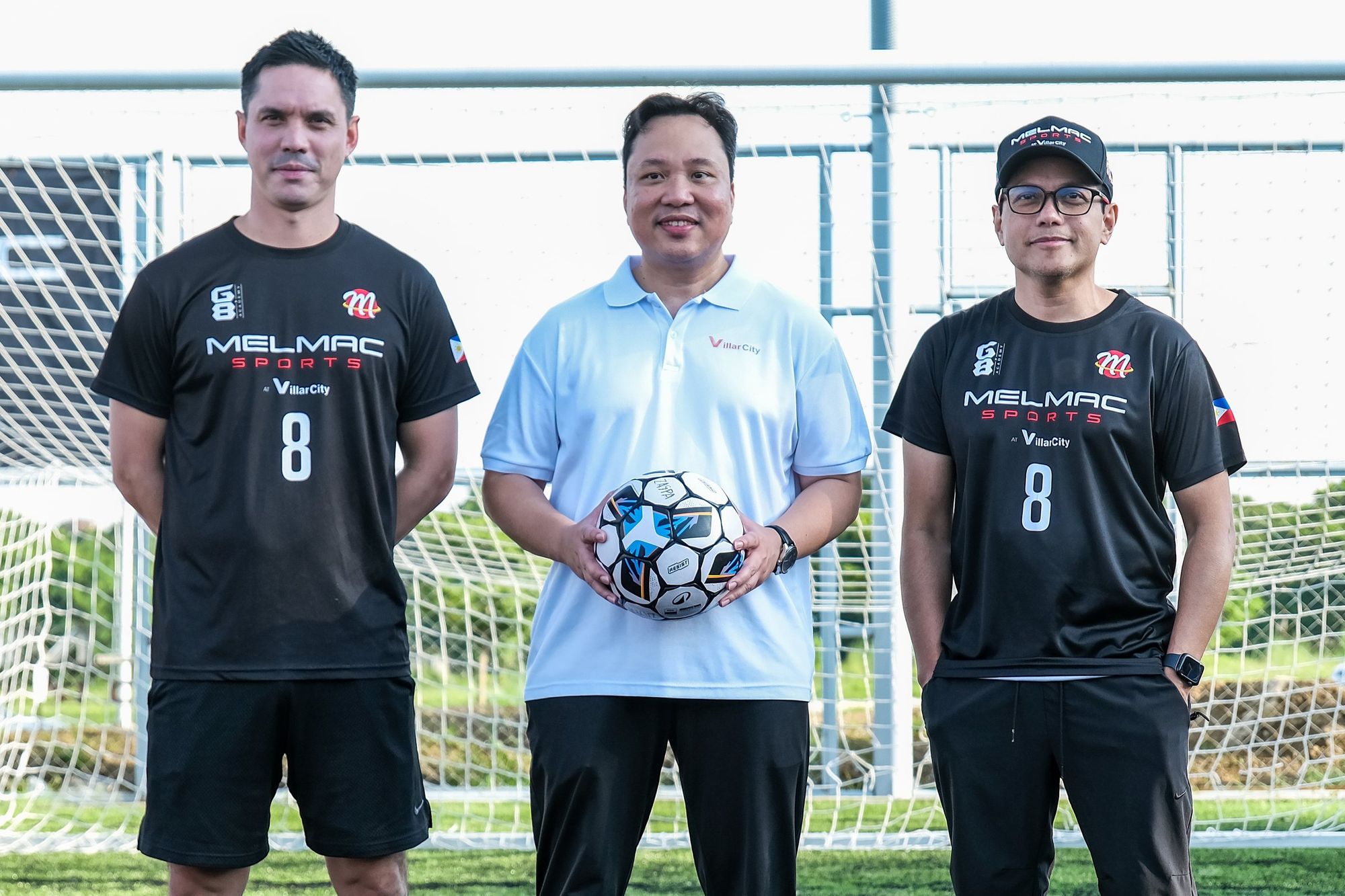 Two men in black Melmac number 8 jerseys and a guest holding a football in front of a goal at Villar City