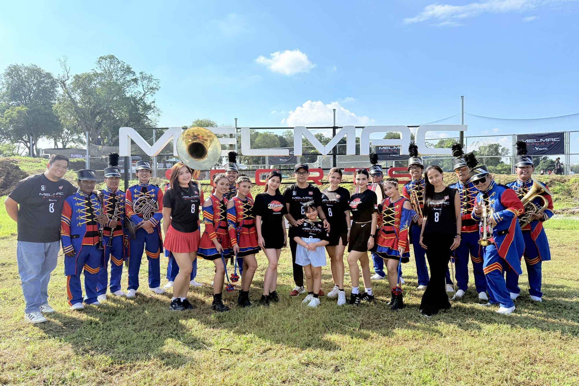 A large group with the marching band in front of the Melmac Sports sign at Villar City