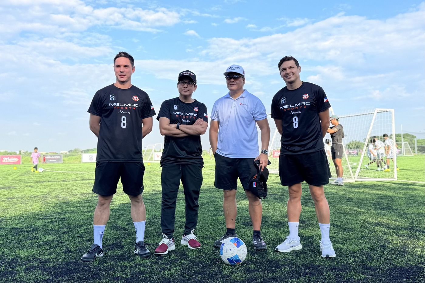 Three men in black Melmac shirts and a guest posing on the pitch at Villar City