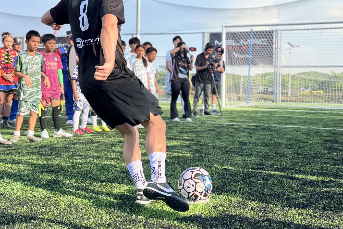 A player in a black number 8 jersey lining up a kick as children watch from the sideline at Villar City