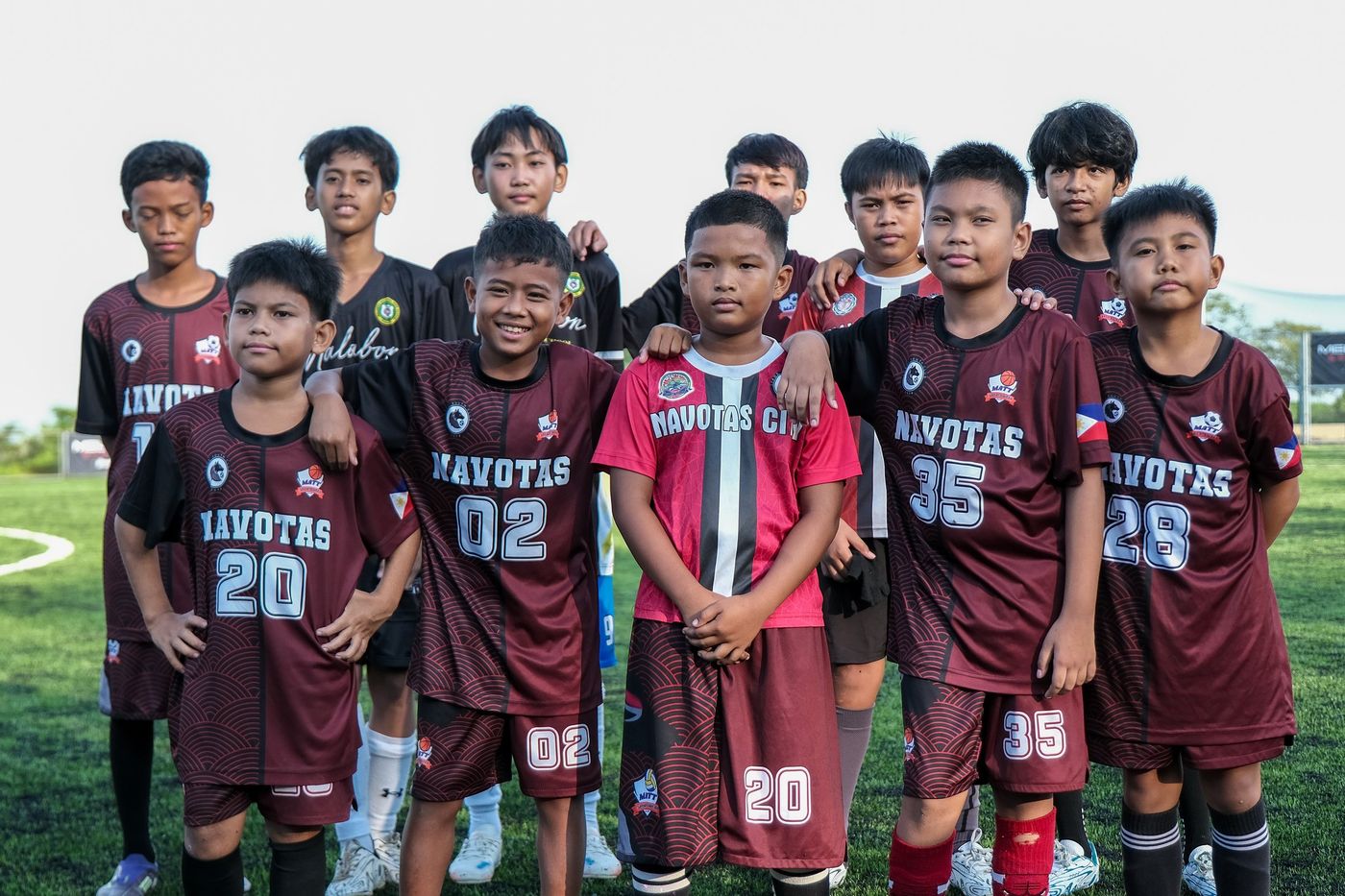 A youth team in maroon kits posing together on the pitch at Villar City